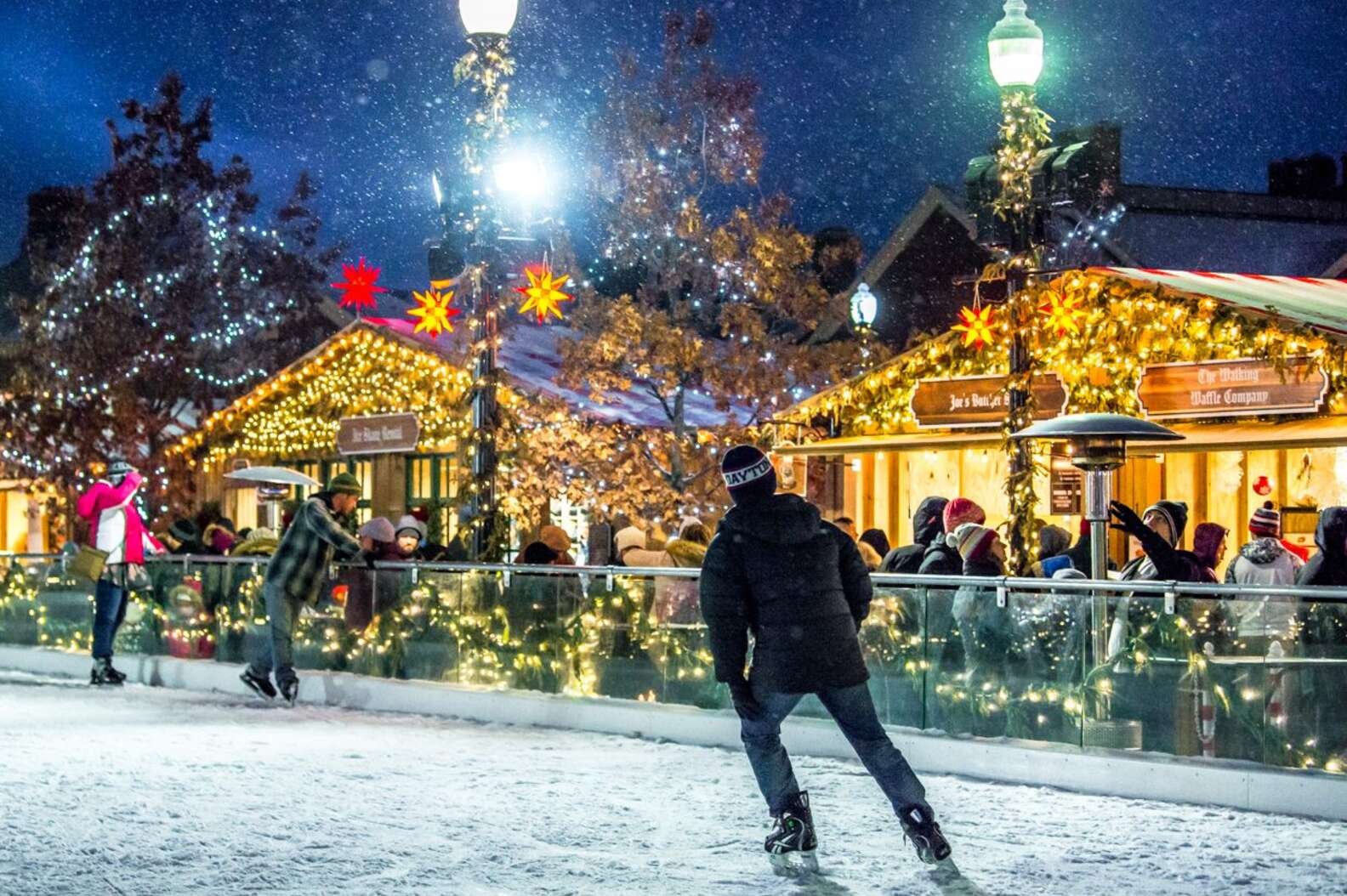 Ice skaters glide on a snowy rink at night beside string-lit wooden market stalls and glowing star ornaments as snow falls