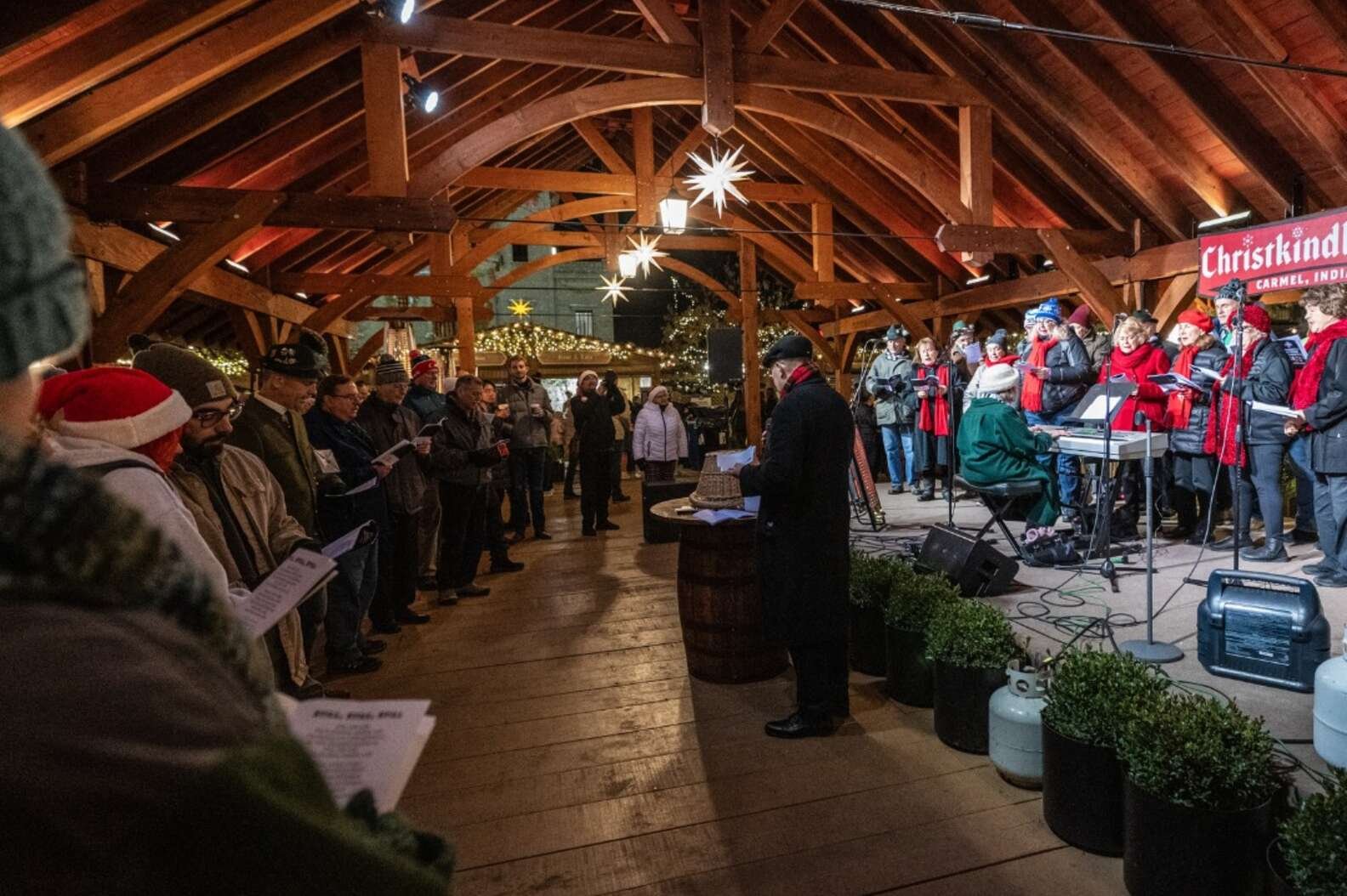 Two caroling groups in winter coats sing under a timber-frame pavilion with illuminated Moravian stars and a Christkindl banner overhead
