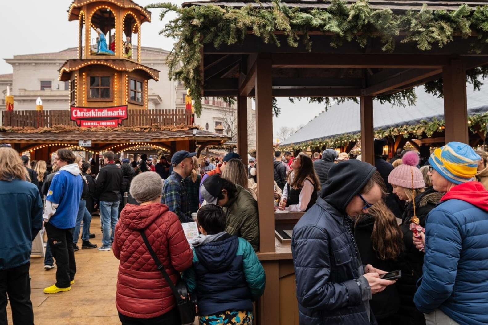 Bundled crowds browse wooden vendor stalls at Carmel's Christkindlmarkt in overcast daylight beneath a glowing Nativity tower